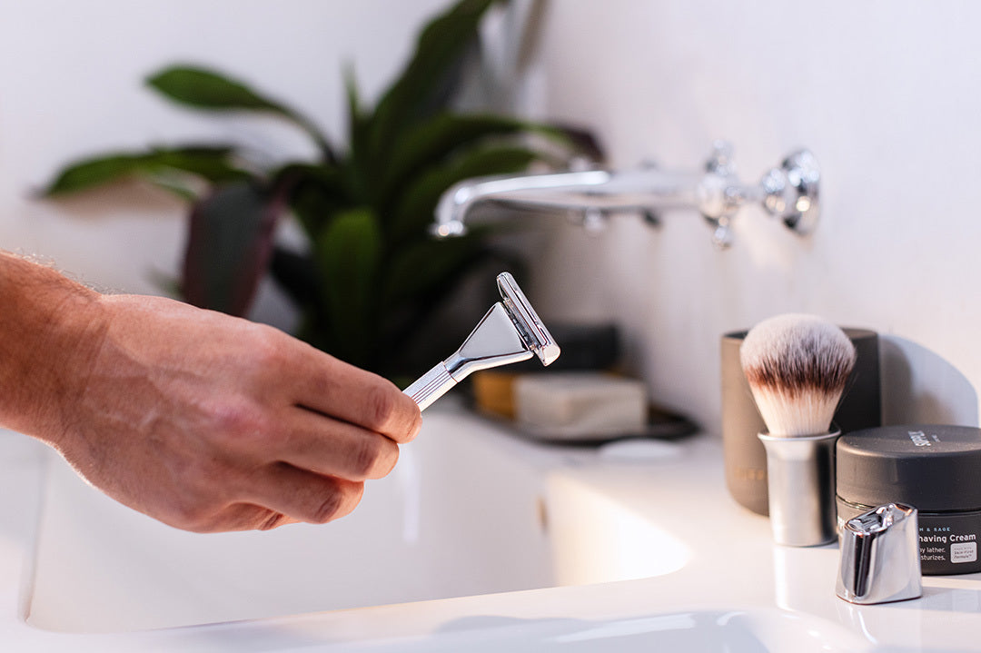 A lifestyle image in a bathroom with someone holding a single edge max razor with the accessories around the sink. 